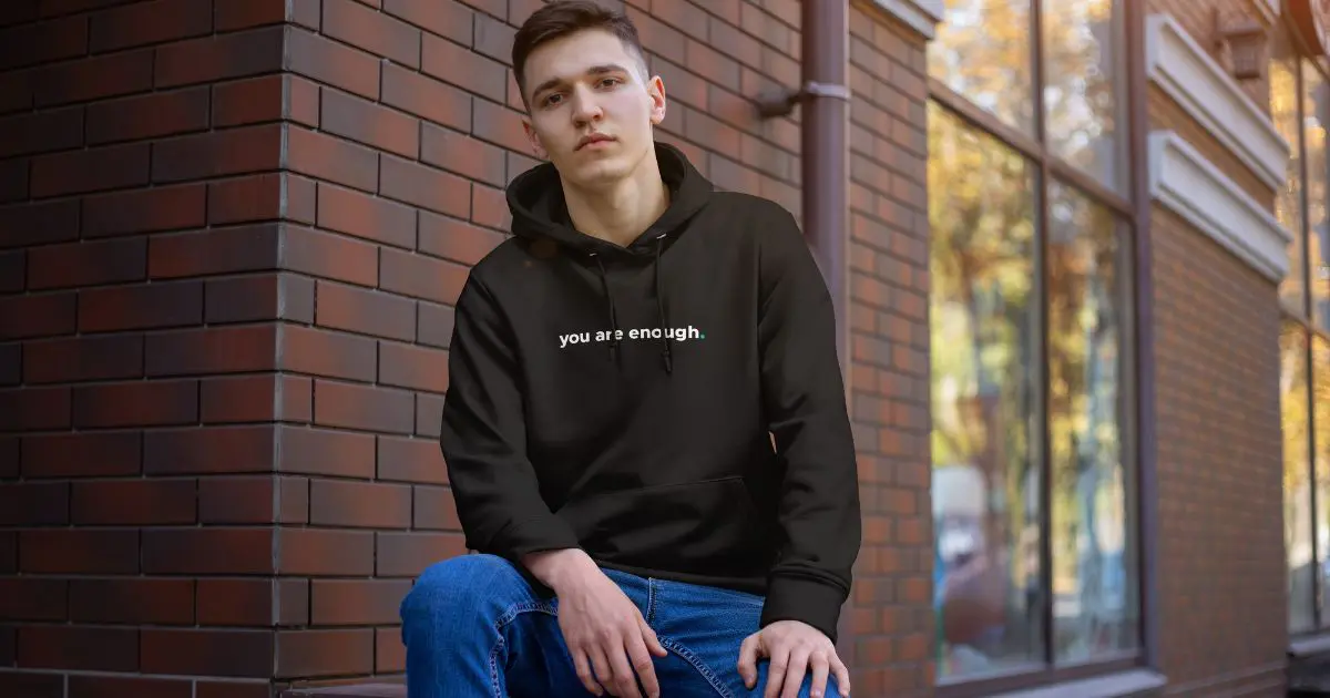 Man wearing a black “you are enough.” statement hoodie while sitting by a brick wall in an urban street setting.